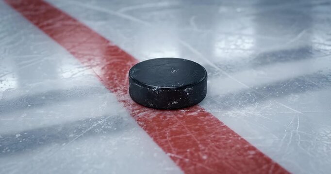 A close-up shot of a black hockey puck resting on a frozen ice rink, with a prominent red line visible across the surface.