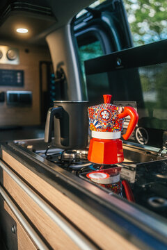 Vibrant red moka pot espresso maker on a gas stove inside a camper van kitchen with forest view, capturing the warmth of a slow morning coffee ritual on the road