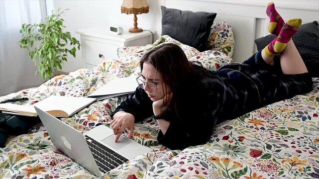Focused teenager studying with open laptop surrounded by books and notes on cozy bed in evening light, comfortable home learning environment and remote education workspace for self directed academic