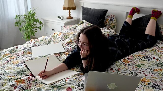 Teen girl sitting on bed with pen and open laptop preparing assignments for school in evening, modern student combining digital and traditional learning tools for remote education and homework