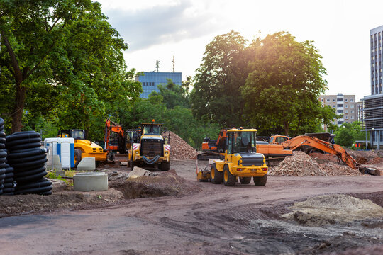 Urban construction site with excavator and dump truck during sunrise near modern buildings and green trees. Heavy machinery works on groundwork and infrastructure development in growing city district
