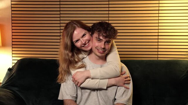 This depicts young couple love and affection, with a happy woman hugging a man from behind while they sit on a sofa. The scene conveys relationship goals and intimacy, projects focusing on romantic re