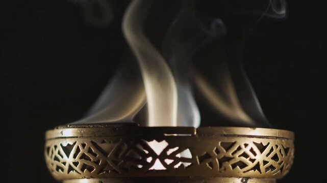 Emitting brass incense burner creating smoke from warm glow in studio, curls widening into ribbons