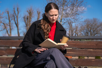 Obraz premium Young woman on a wooden park bench reading a book and holding a takeaway coffee, enjoying a quiet, thoughtful moment outdoors in cool weather among bare trees and sunlight