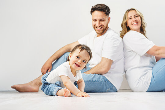 A cheerful family with a happy, crawling baby. The parents are smiling and looking lovingly at their daughter. Everyone is wearing casual white tops and jeans.