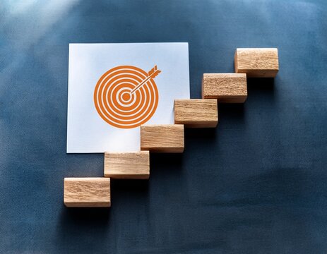 a clear visual metaphor of achieving business goals and continuous progress represented by wooden steps with targets leading to a final target on paper