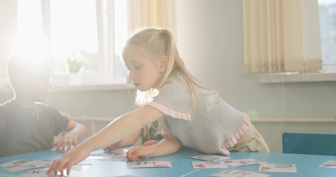 Young children playing an educational card game together at a classroom table, collaborating and learning in warm sunlight by large windows