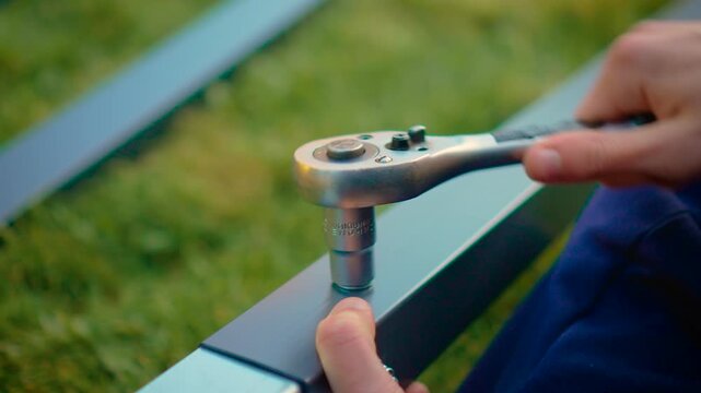 Close view hand turning ratchet tool securing bolt on rectangular metal frame. Mechanical installation process with equipment and aligned parts on grass background. Home repair theme with careful