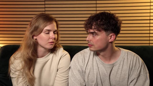 Relationship conflict and emotional distance. A young couple sits apart on a sofa, looking away from each other, conveying a sense of misunderstanding and unresolved issues. This footage is editorial 