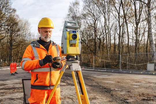 Worker checks equipment while using a total station at a construction site in the afternoon near trees