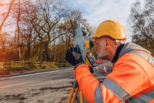 Construction worker uses surveying equipment in a wooded area to measure and plan roadwork during the day