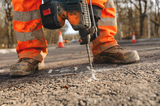 Worker drills into asphalt at a road construction site in the late afternoon near a busy area with traffic