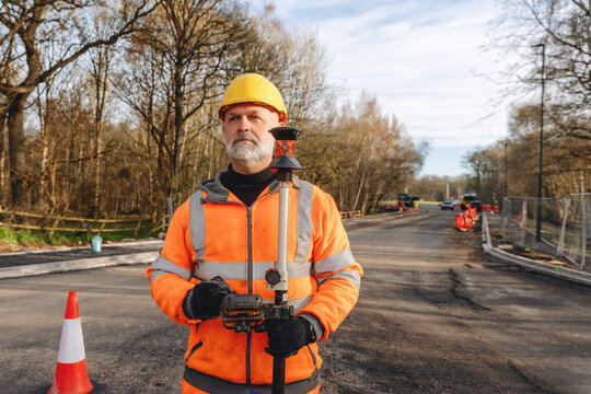 Construction worker stands on road site wearing safety gear and holding equipment in woodlands during daytime