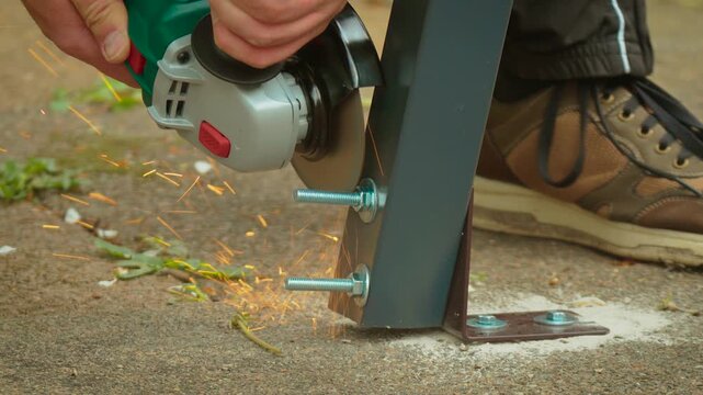 Electric grinder slicing through threaded rod on mounted metal frame with intense spark stream. Hand controlling power tool while removing excess fastener length for proper assembly. Construction