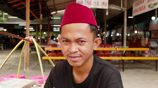 Portrait of young indonesian street vendor in songkok hat selling local food at street food stall in Jakarta market, Indonesia