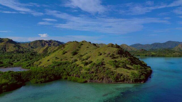 Komodo island landscape with lush hills, clear ocean, and a simple wooden pier extending to shore. Amazing aerial view descending panorama drone
