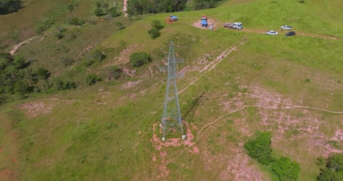 Steel lattice transmission tower surrounded by pasture hills, maintenance vehicles and utility work area near Santo Ant&ocirc;nio do Monte, Minas Gerais, Brazil, drone orbit aerial shot.