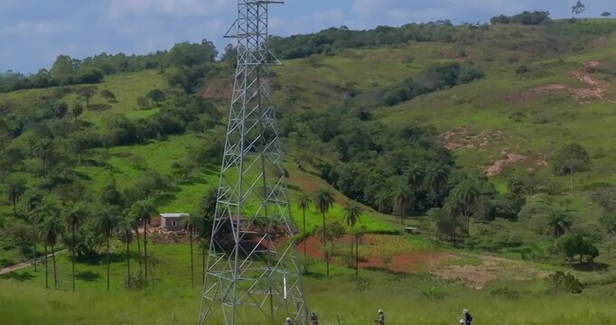 Utility maintenance team working beneath a steel lattice transmission tower near Santo Ant&ocirc;nio do Monte, Minas Gerais, Brazil surrounded by pasture hills and fragmented rural vegetation aerial descent