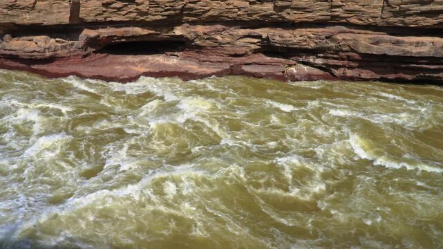Detailed high-angle shot of the turbulent Yellow River current rushing past a red sandstone cliff at Hukou. Shows the heavy sediment load and churning white foam of the rapids.