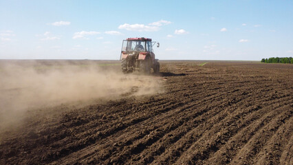 Fototapeta premium drone shot capturing a red tractor pulling a harrow across a vast, dark-soiled field. Dust trails behind the machinery as it prepares the land for the planting season under a clear blue sky.
