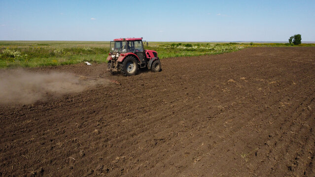 drone shot capturing a red tractor pulling a harrow across a vast, dark-soiled field. Dust trails behind the machinery as it prepares the land for the planting season under a clear blue sky.