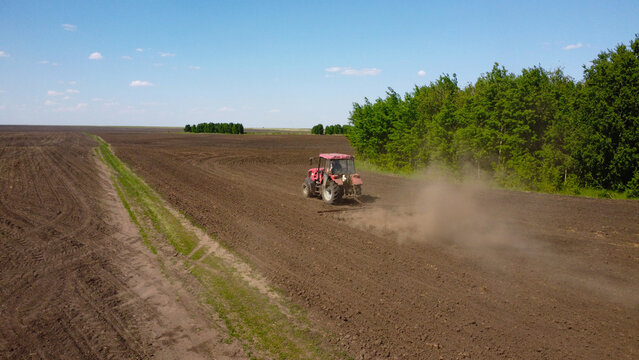 drone shot capturing a red tractor pulling a harrow across a vast, dark-soiled field. Dust trails behind the machinery as it prepares the land for the planting season under a clear blue sky.