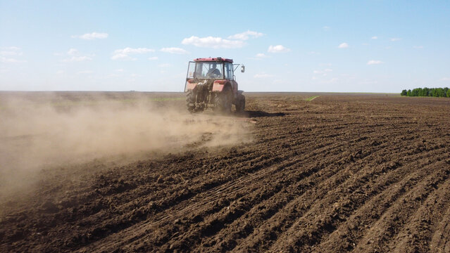 drone shot capturing a red tractor pulling a harrow across a vast, dark-soiled field. Dust trails behind the machinery as it prepares the land for the planting season under a clear blue sky.