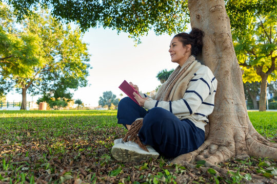 Woman enjoying quiet time reading a book under a large tree in a sunny park, finding peace and relaxation outdoors