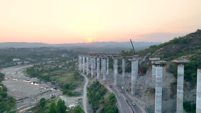 Massive concrete pillars of incomplete highway flyover in India. Sweeping aerial drone shot showcasing the stark contrast between towering, modern concrete bridge pillars and the natural river valley.