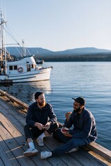 Fototapeta premium Two men laughing and eating on a wooden dock near a fishing boat for lifestyle banner and coastal background