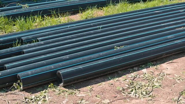 Long black plastic pipes are arranged in neat rows on grassy ground in an outdoor area. The setup appears to be for a construction project involving plumbing or irrigation work