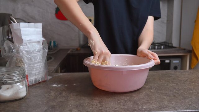 Unrecognizable woman wearing a dark blue t-shirt kneading sticky dough in a pink plastic bowl on a kitchen counter, showing the initial stages of preparing homemade bread or pastry