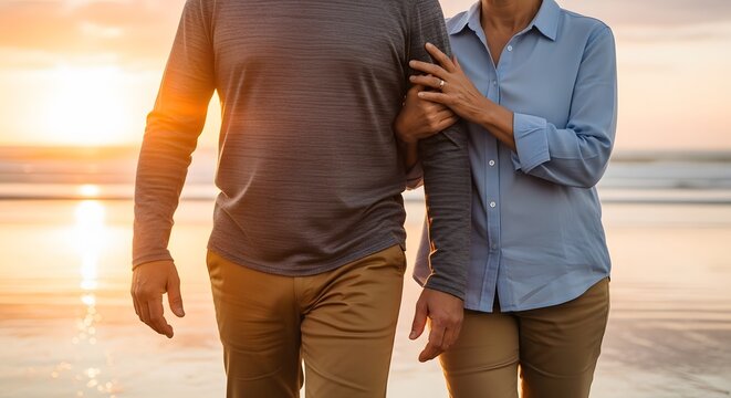 Close up of senior couple linking arms while walking together on beach at golden sunset
