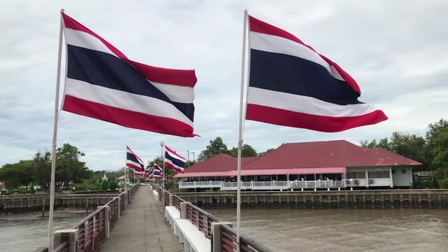 Rows of Thai flags flutter on poles lining a pedestrian pier beside a Chao Phraya river, slow motion