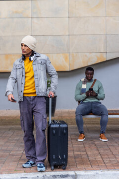 Diverse men standing, holding black suitcase, seated man checking phone at transit stop, copy space