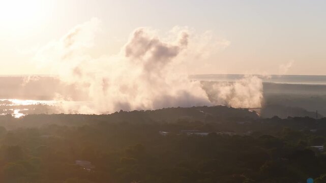 Aerial sunset view over Victoria Falls, Zimbabwe, with spray rising, the arched Victoria Falls Bridge at right, warm light, long shadows, and high atmospheric haze.