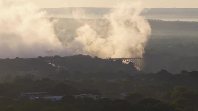 Aerial sunset view shows Victoria Falls spray billowing and drifting in Zimbabwe. Victoria Falls Bridge spans the chasm as a village and trees sit in the foreground.