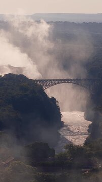 Aerial sunset shows Victoria Falls, mist plumes, and Victoria Falls Bridge on the Zimbabwe and Zambia border, with a Zimbabwe village, warm backlight, and soft haze.