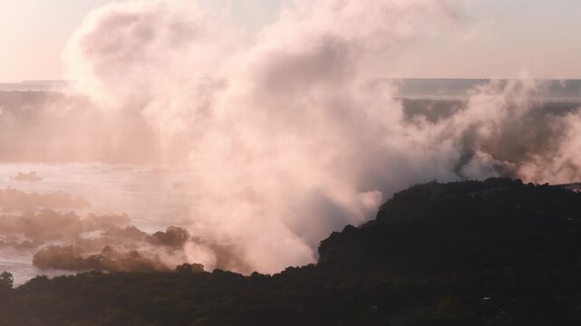 Aerial sunset view of Victoria Falls in Zimbabwe shows the Zambezi River, a rising spray plume that drifts and shifts, shadowed cliffs, islets, and golden hour light.