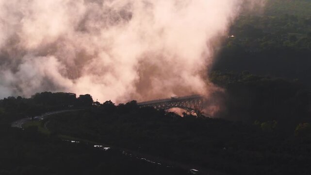 Aerial view shows Victoria Falls Bridge spanning the gorge at sunset, mist rises and glows, rainforest and road sit in silhouette, tiny cars and people visible.