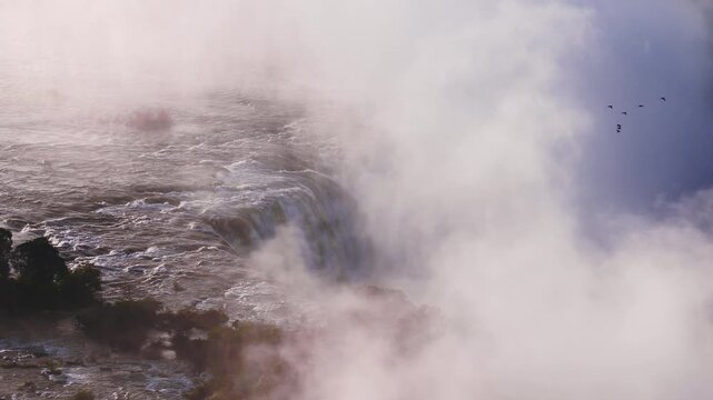 Aerial view shows Victoria Falls as water tumbles over the lip, mist rises, rapids churn, and birds arc through vapor near tree covered islands at sunset.