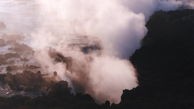 Aerial dusk scene shows Victoria Falls in Zimbabwe as spray plumes rise, mist drifts, and the Zambezi roils past rocky islets, forested cliffs, and a rim road in warm light.