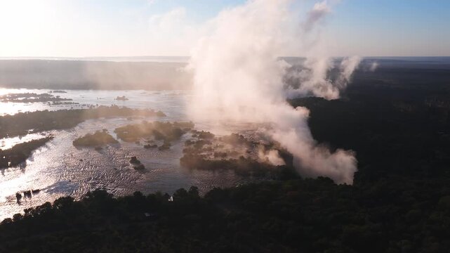 Aerial view passes Victoria Falls in Zimbabwe as mist plumes rise, braided channels flow, islands dot the Zambezi, and warm late afternoon light reflects on water.