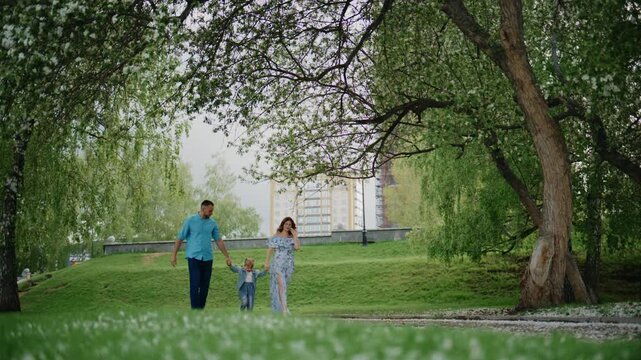 Family walks through green park under blooming trees. Adults hold child's hands, showing care and joy. Scene captures peaceful family time outdoors. Ideal for parenting, nature, or lifestyle content