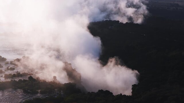 Aerial sunset view of Victoria Falls on the Zimbabwe Zambia border. Spray rises, expands, and drifts downstream as golden light backlights islands and silhouettes.