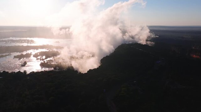 Aerial pan at golden hour shows Victoria Falls gorge and spray, island rapids, forested banks, glittering water, and drifting mist, with village clearings in Zimbabwe.