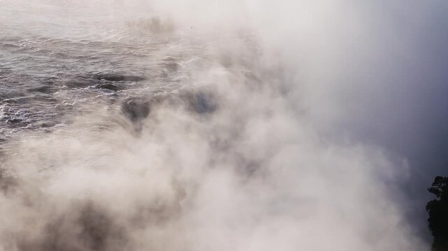 Aerial view shows Victoria Falls on the Zimbabwean side at sunset. Thick mist drifts, spray rises, birds flit, treeline silhouettes, warm backlight, foamy water churns.