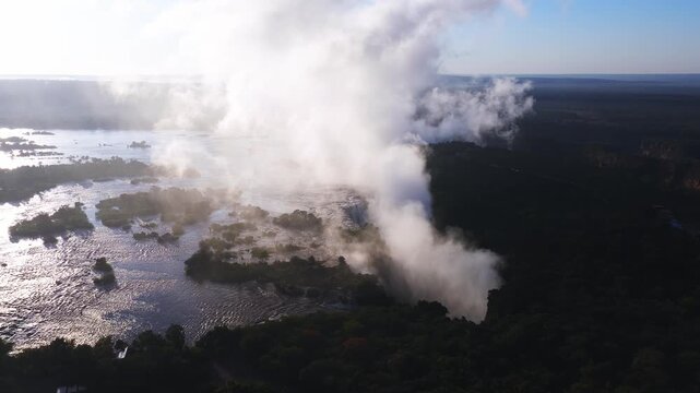 Aerial wide shot shows Victoria Falls in Zimbabwe at sunset. Spray rises, water thunders into a dark gorge, mist drifts, river islands and riverside buildings appear.