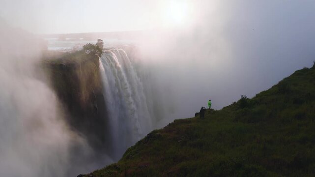 Aerial view shows Victoria Falls in Zimbabwe as water plunges into mist. A lone tree marks the lip. Two figures in rain gear photograph the scene at sunset.
