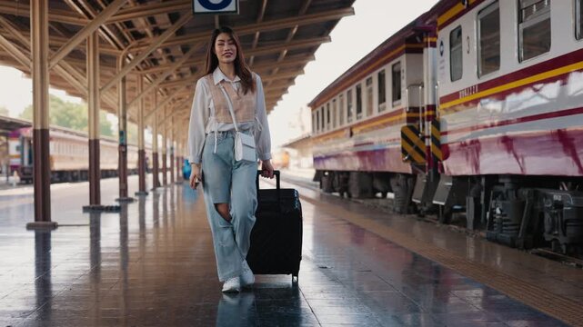Portrait of thoughtful Asian female traveller with smartphone and luggage walking along railroad platform. Brunette woman with rolling suitcase boards train at transport station